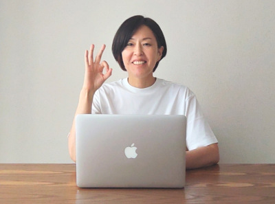 白いTシャツの女性がパソコンの前で笑顔でOKサインをしている画像